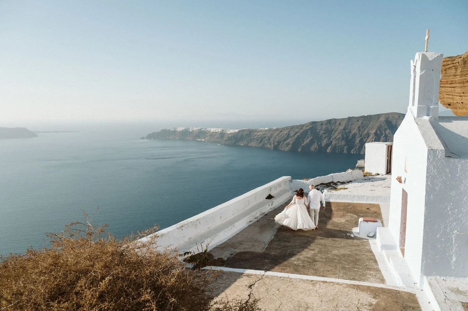 A bride and groom walking hand in hand on a terrace overlooking the ocean and cliffs in Santorini, Greece, during their wedding.
