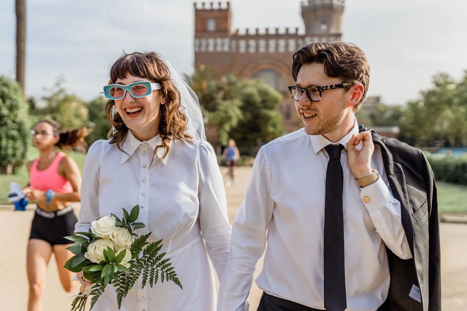 A smiling woman dressed in white holding a bouquet of white roses and greenery, walking outdoors with a man in glasses and a black tie, both dressed in white shirts. In the background, a woman in a pink top and sunglasses is seen jogging, and a histo