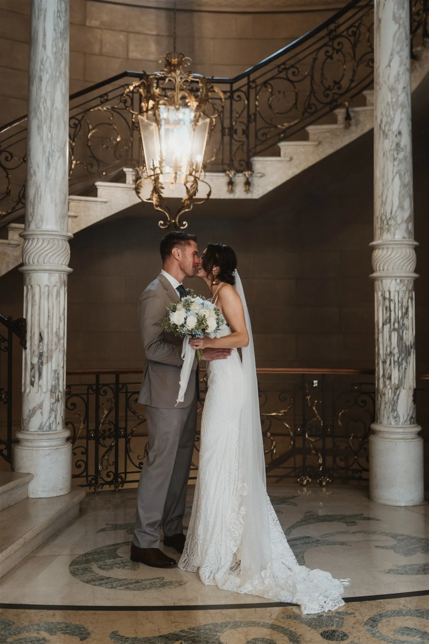 A bride and groom are standing close together indoors, embracing and about to kiss, underneath a grand chandelier and ornate staircase railing.