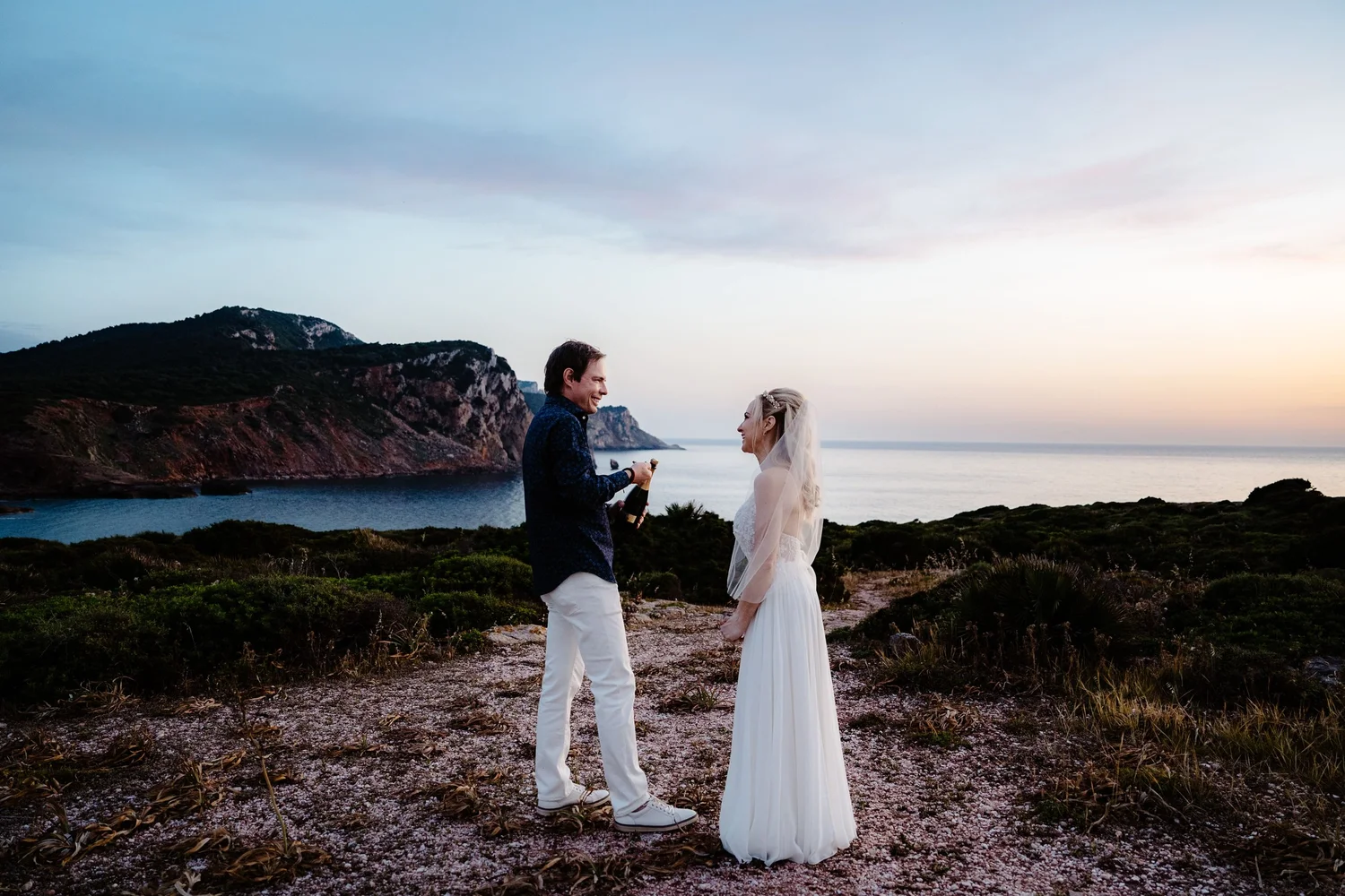 A couple dressed in wedding attire standing outdoors near the coastline at sunset. The man is proposing or giving a gift to the woman, who is smiling back at him. There are mountains and the ocean in the background.