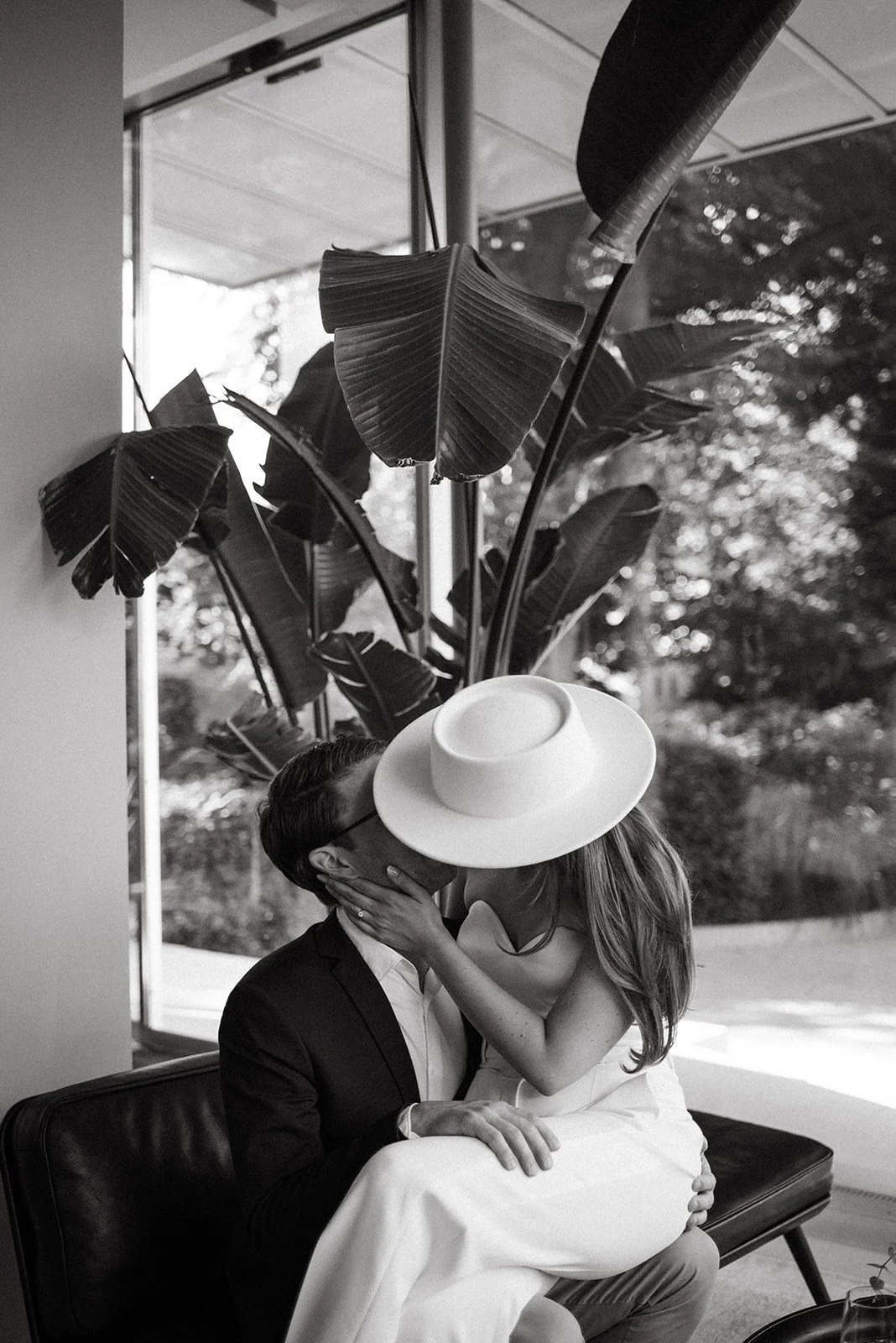 A couple kissing indoors, the woman wearing a large hat and a strapless dress, the man in a suit, with large tropical plants in the background and sunlight coming through windows.