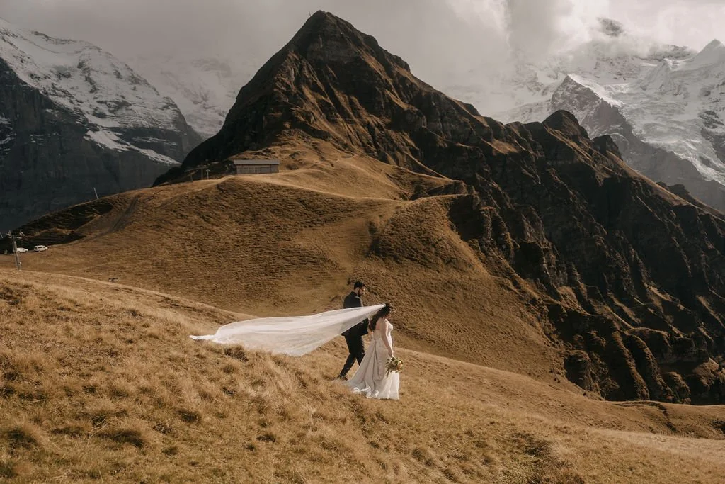 A bride and groom standing on a grassy hillside with mountain peaks and snow-capped summits in the background, with the bride holding a bouquet of flowers and the groom standing behind her, the bride's veil flowing in the wind.