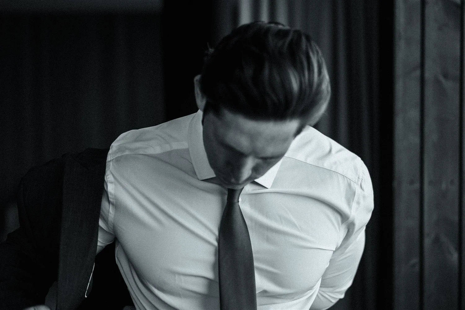 A man in a white dress shirt and tie, with glasses, looking down, in a dark room with curtains.