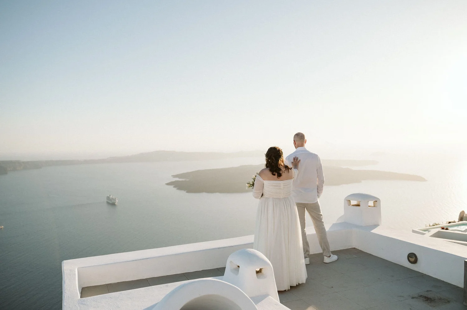 A bride and groom standing on a white terrace overlooking the sea at sunset or sunrise, with the bride wearing a long white dress and the groom in light-colored clothing, facing away from the camera.