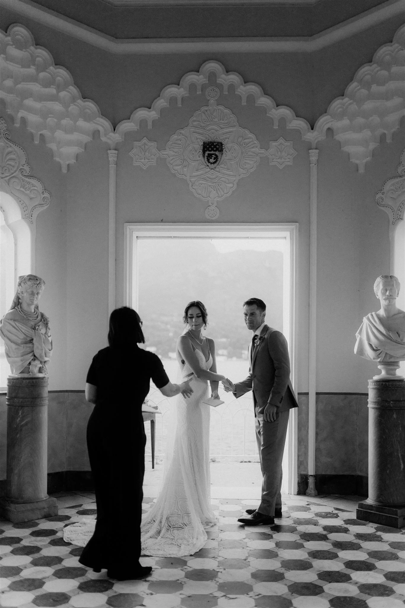 A black and white photo of a wedding ceremony taking place indoors. There is a bride and groom facing each other and holding hands. A person, possibly the officiant, is standing in front of them. Two bust sculptures on pedestals are on either side of