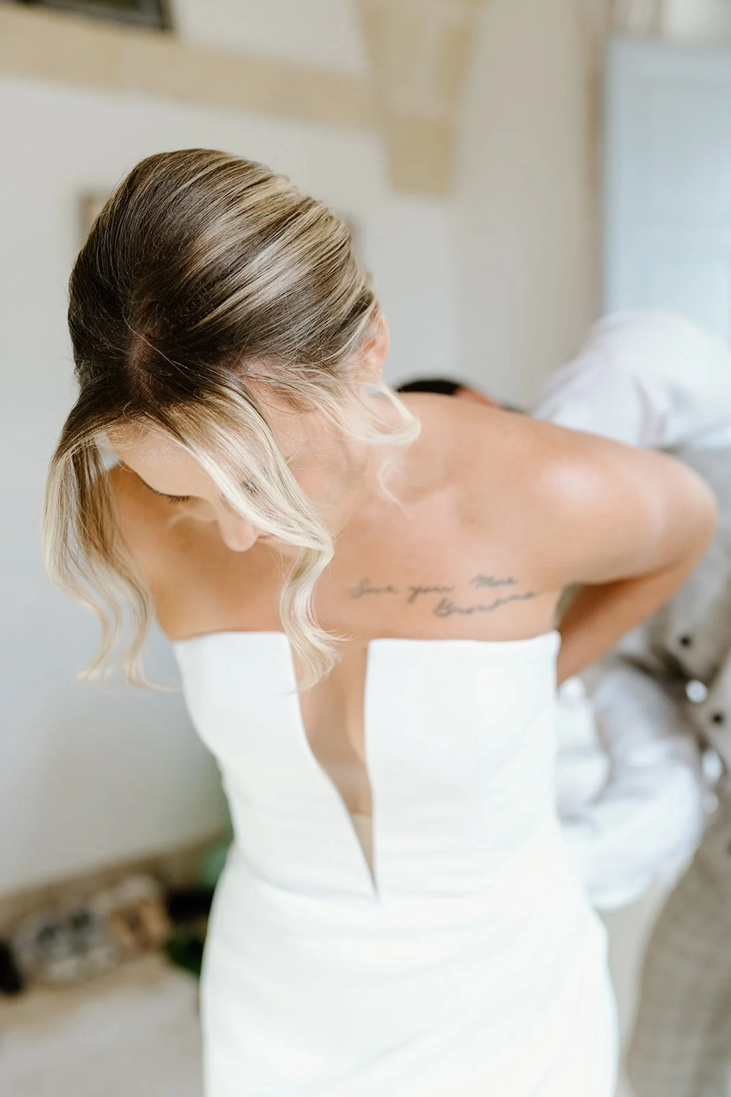 Woman in a white dress with blonde hair styled in loose waves, showing her back and tattooed shoulder, in a softly lit room.