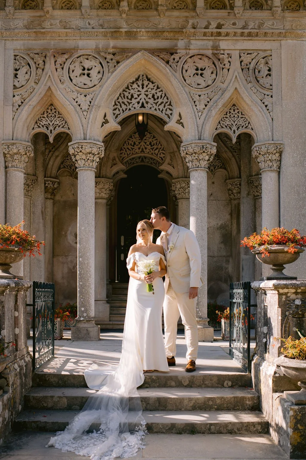 A newlywed couple dressed in wedding attire standing on the steps of an ornate stone building with gothic arches, surrounded by planters with orange flowers. The bride is holding a bouquet, and the groom is kissing her on the forehead.