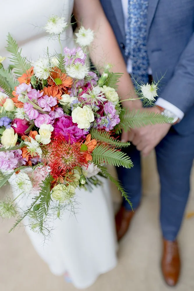 Close-up of a colorful bouquet of flowers held by a bride, with partial view of a groom standing nearby in a suit.
