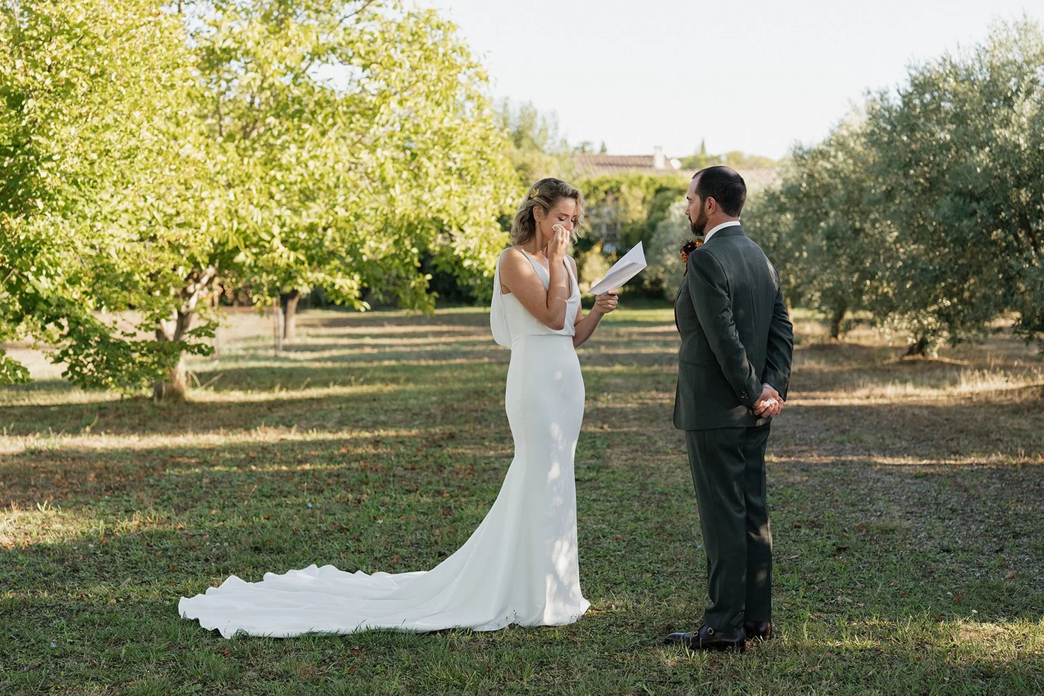 A bride in a white wedding dress crying as she recites vows during an outdoor wedding ceremony, standing opposite a groom dressed in a dark suit under a clear sky with lush green trees in the background.