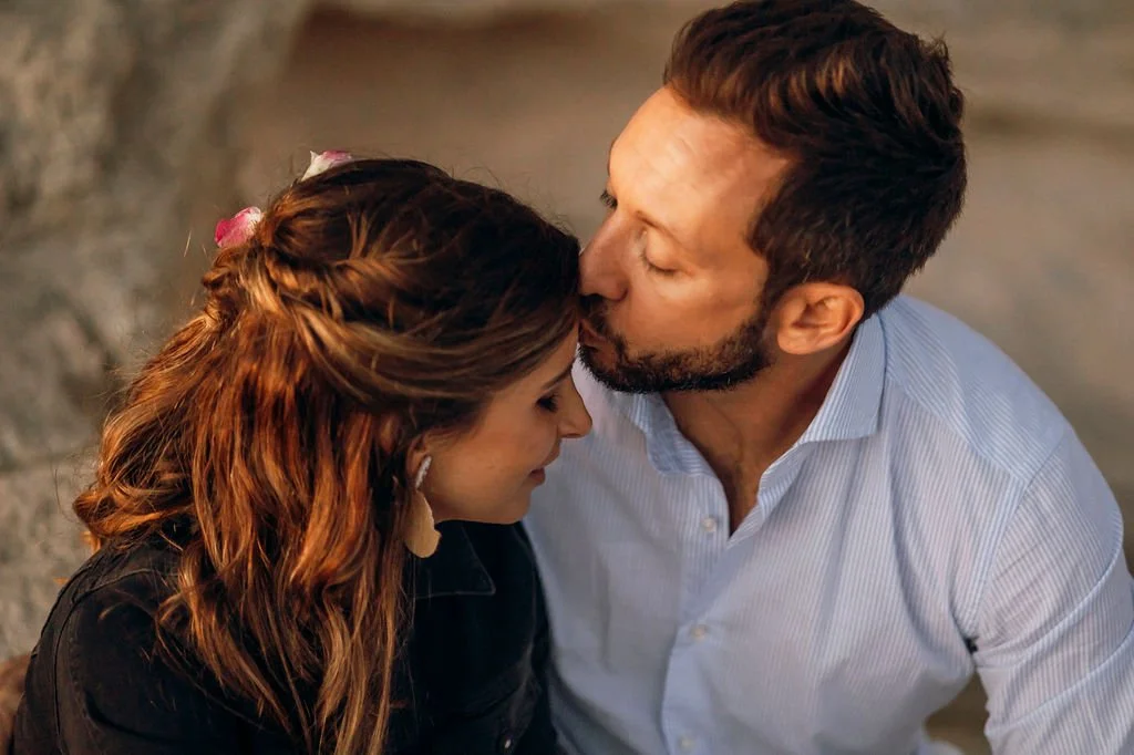 A couple sharing an intimate moment, with the man kissing the woman's forehead as they sit close together.