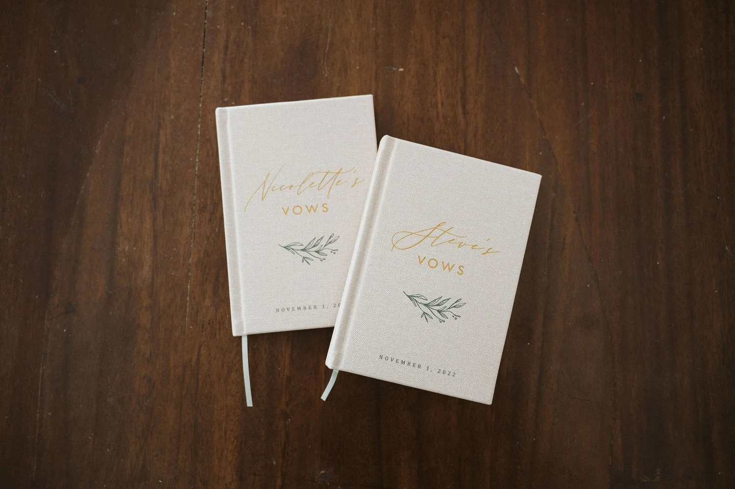 Two white vow books with gold and green lettering and design, placed on a wooden surface.