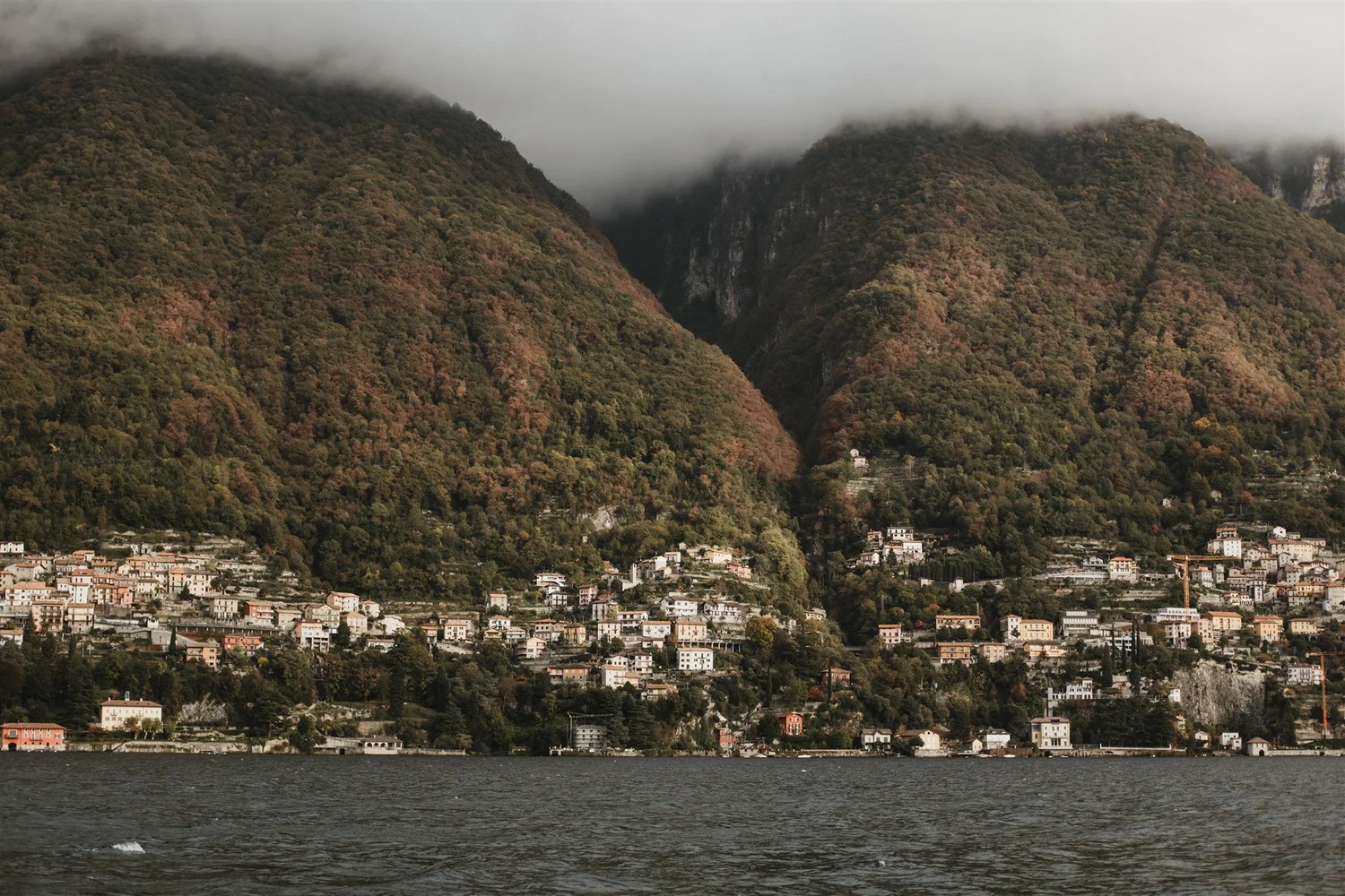 View of a small town at the base of green, forested mountains partially covered with fog, with a body of water in the foreground.