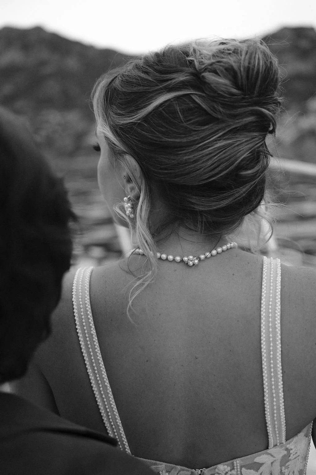 Black and white photo of a woman with an elegant updo hairstyle, wearing pearl earrings and a pearl necklace, seen from behind, outdoors with mountains in the background.