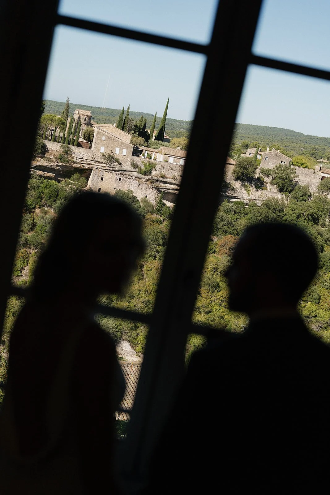 Silhouettes of two people facing each other, seen through window bars, with a scenic view of trees, hills, and buildings in the background.