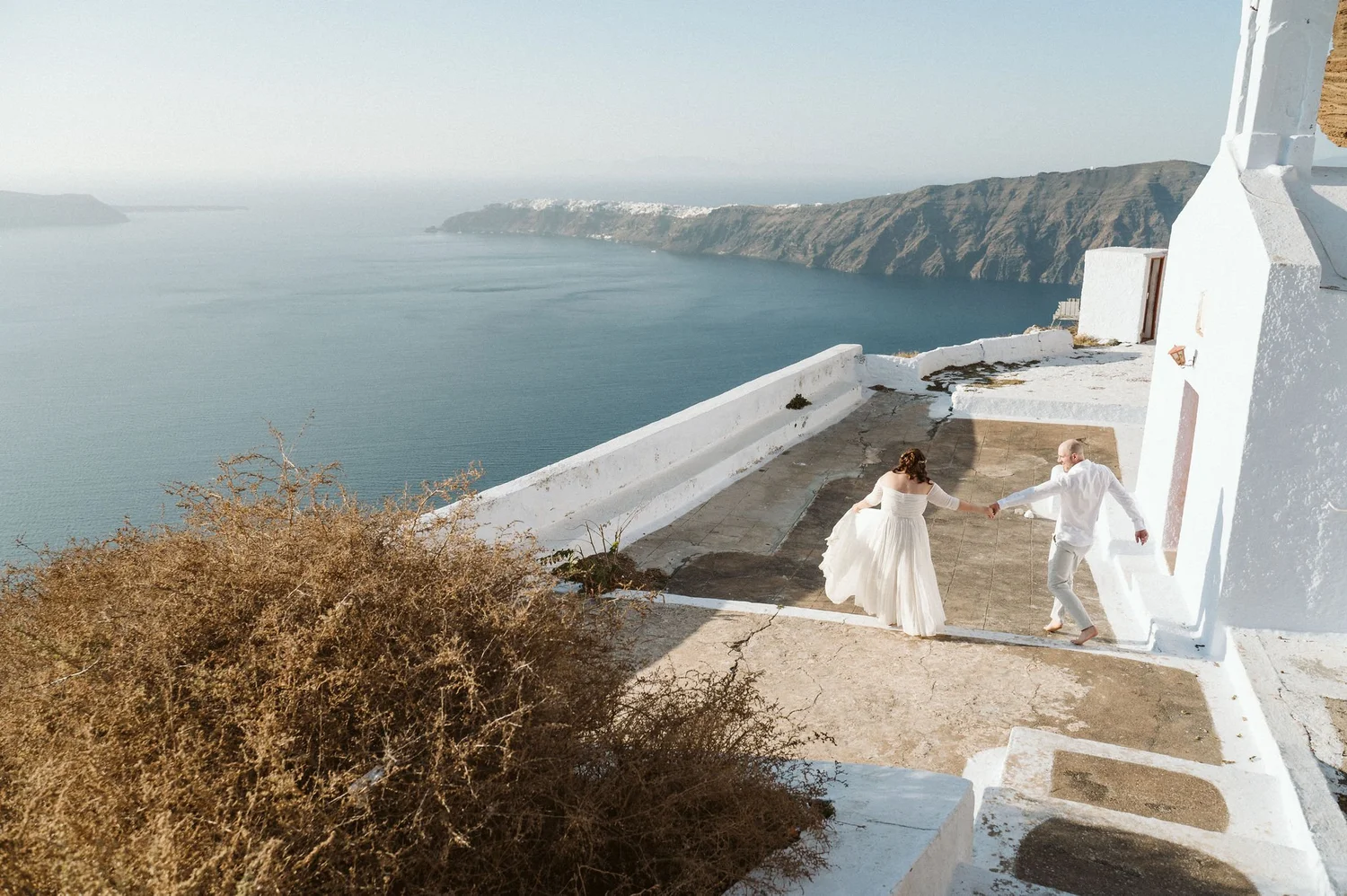 A couple in wedding attire dancing on a small outdoor terrace with a scenic view of the sea and cliffs in the background, with a white building partially visible on the right.