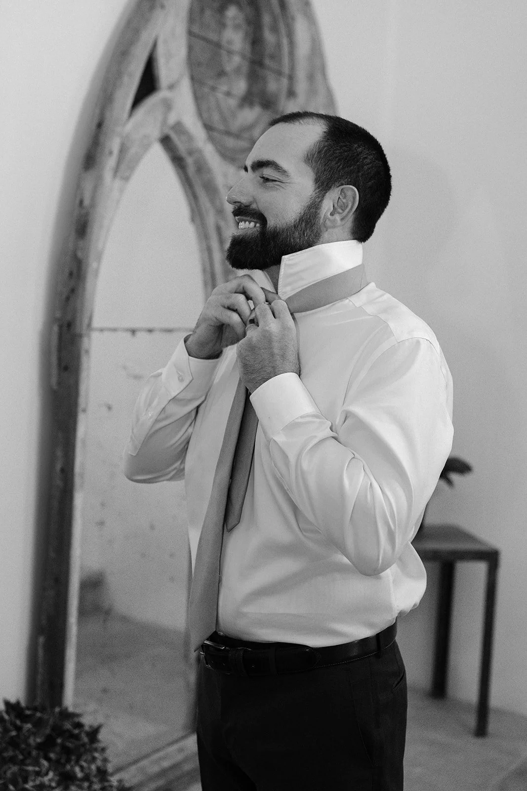 A man with a beard and dark hair smiling while adjusting his tie in front of a mirror.