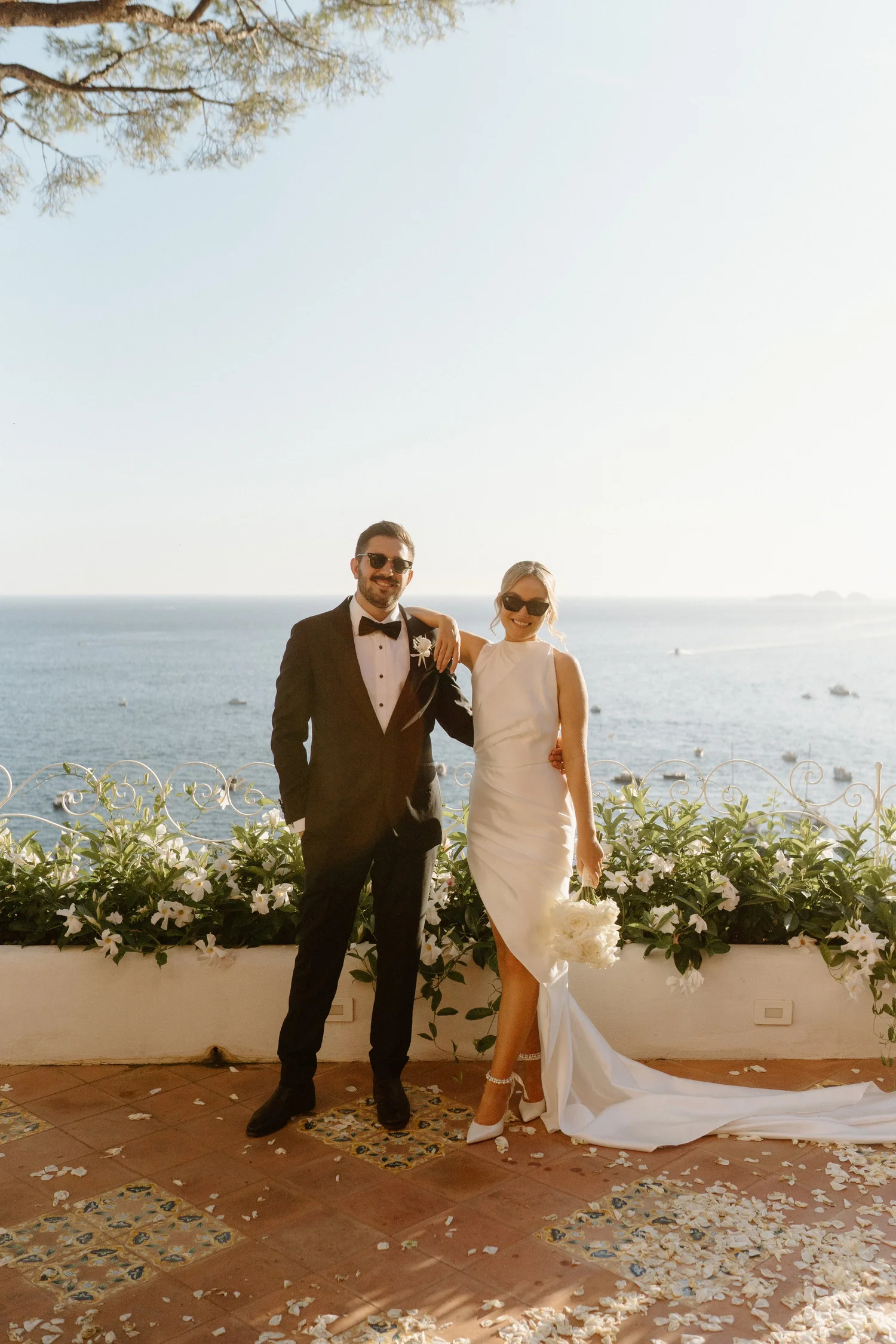 A man and a woman in wedding attire standing outdoors by a railing with a scenic ocean view in the background. The man is wearing a black tuxedo with a bow tie and sunglasses, while the woman is in a white sleeveless wedding gown holding a bouquet an