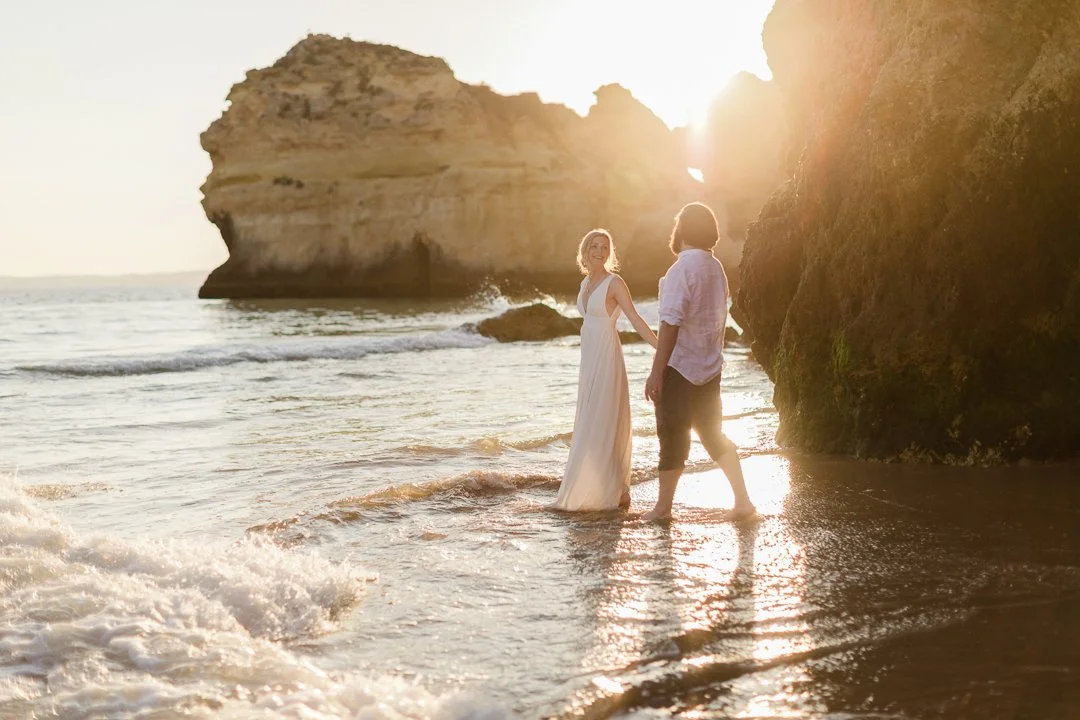 A couple holding hands standing in the shallow surf on a beach at sunset, with rocky cliffs in the background.