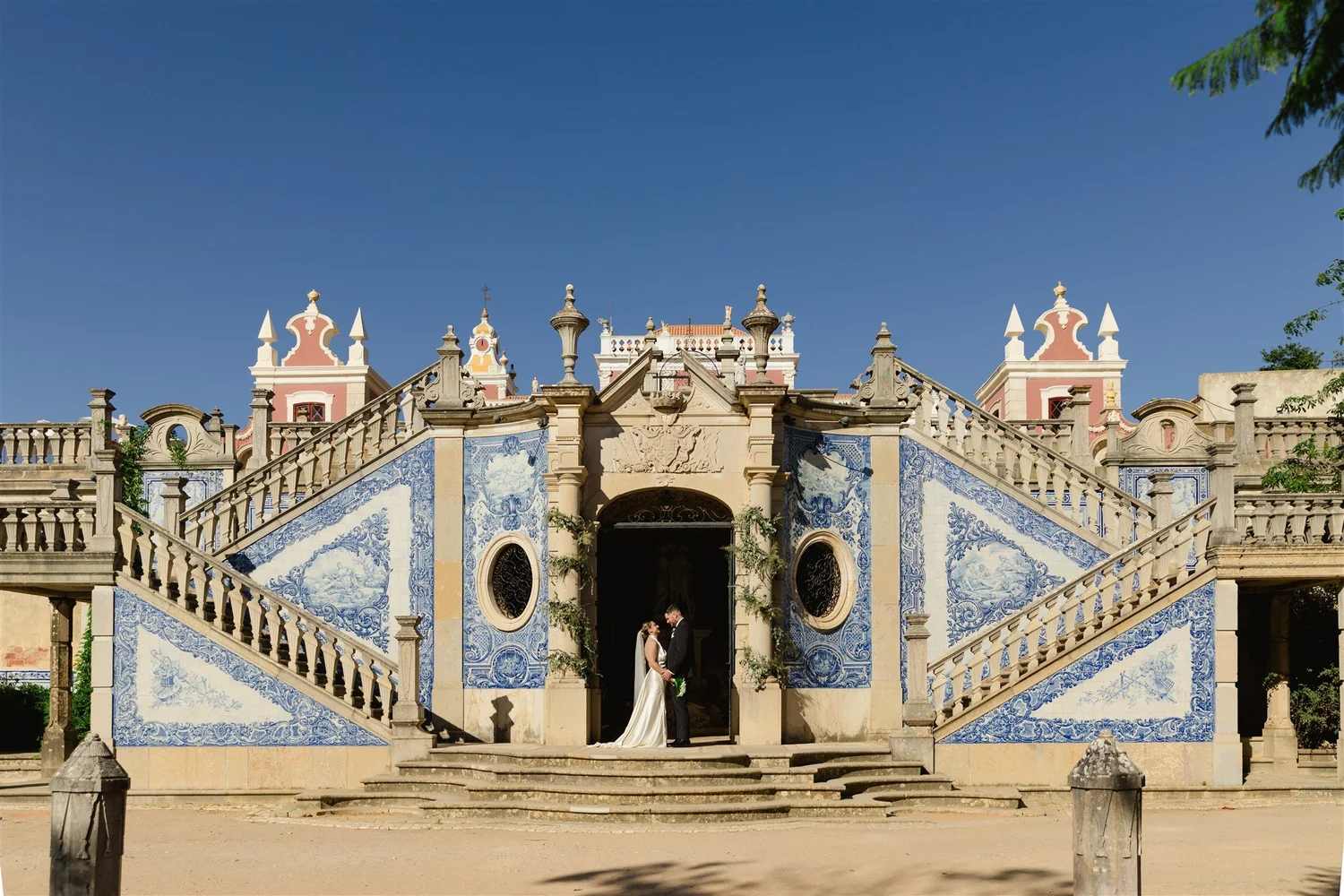 A bride and groom stand under a historic, ornate stone staircase leading to a pastel pink and white castle with detailed blue and white patterned tiles, decorative towers, and a clear blue sky overhead.