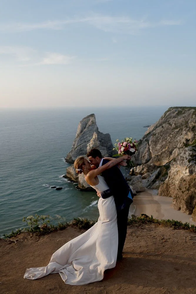A bride and groom kissing on a cliffside overlooking the ocean with rocky formations and a sandy beach in the background, during sunset.