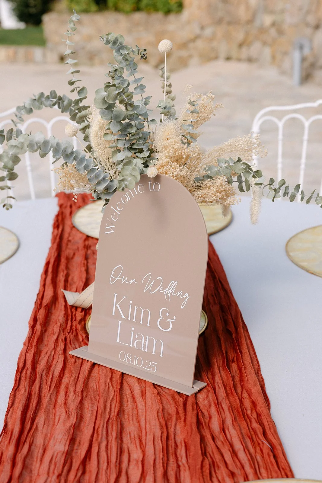 Wedding table centerpiece with a beige welcome sign for Kim and Liam's wedding, dated 08.10.25. The sign is decorated with dried flowers and eucalyptus, placed on a textured red table runner with a white tablecloth.