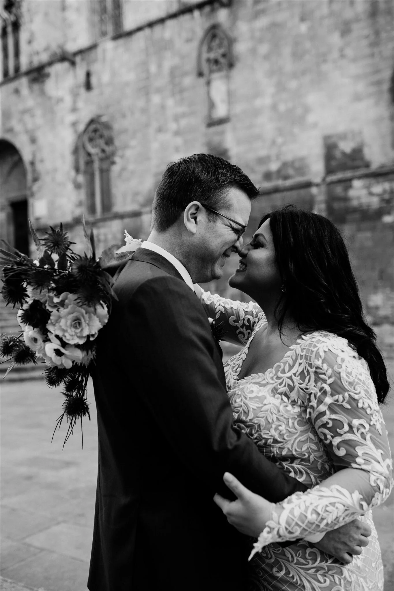 Black and white photo of a happy couple sharing a close moment outdoors, with a brick building in the background. The man is wearing a suit and glasses, holding a bouquet of flowers, and the woman has dark wavy hair and wears a lace dress.