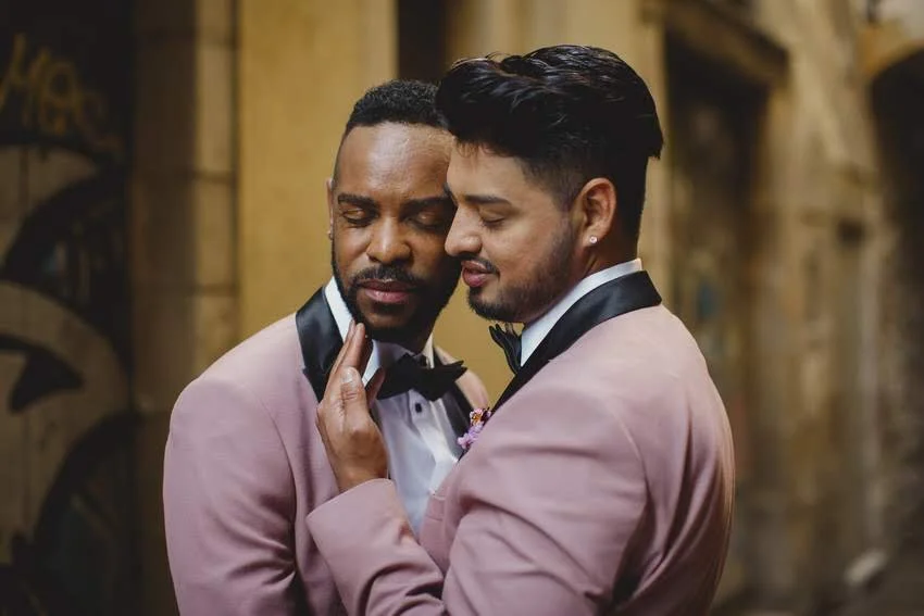 Two men in tuxedos with pink jackets and black bow ties embracing closely in a romantic moment.