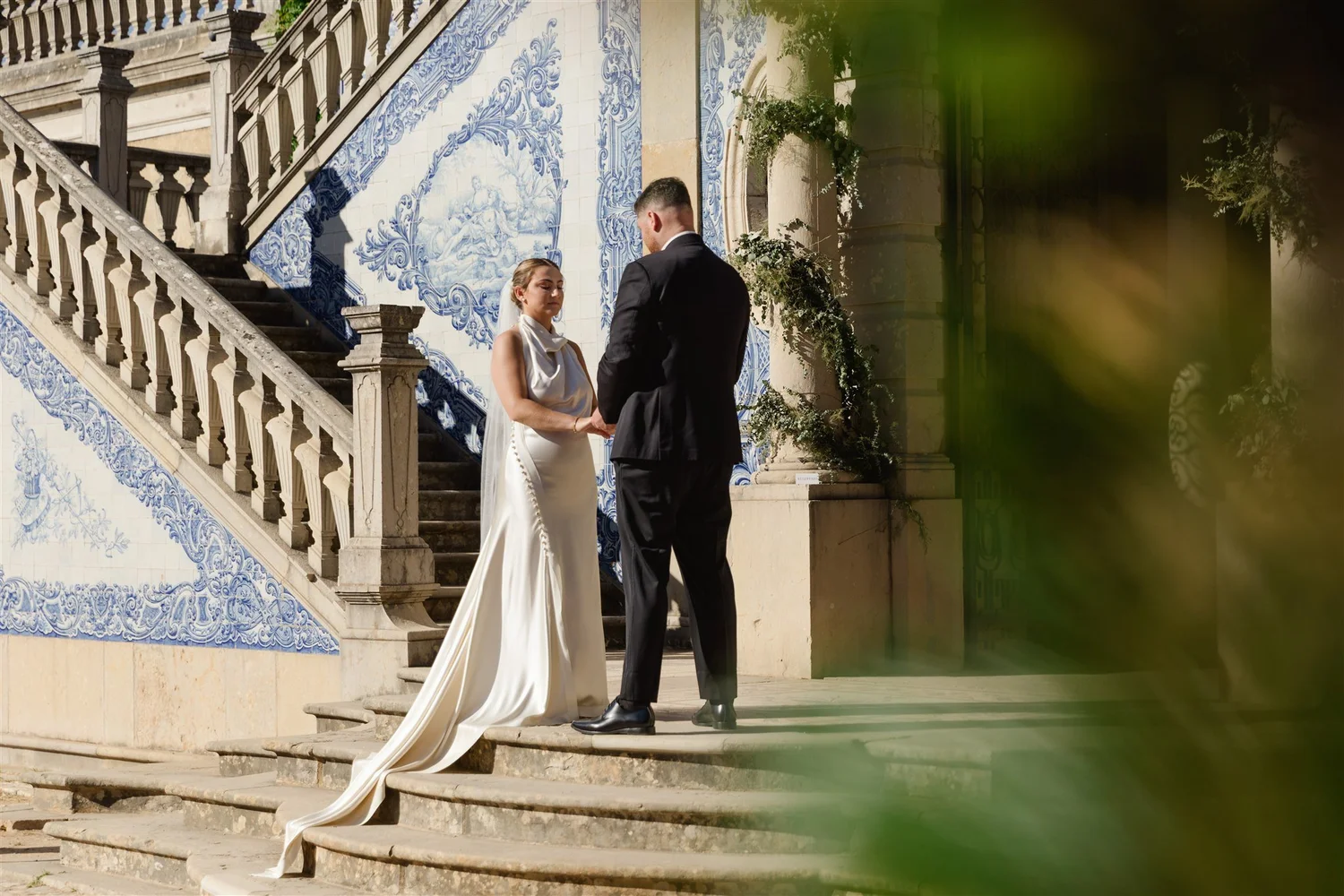 A bride and groom during their wedding ceremony on outdoor steps, with the bride in a white dress and the groom in a black tuxedo, holding hands and standing in front of a decorative blue and white tiled wall.