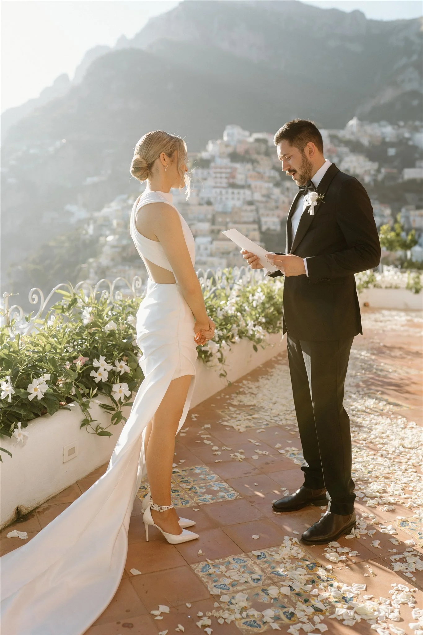 A bride and groom exchanging vows outdoors, standing on a decorated tile with flower petals scattered around, in front of a scenic mountainous background on a sunny day.
