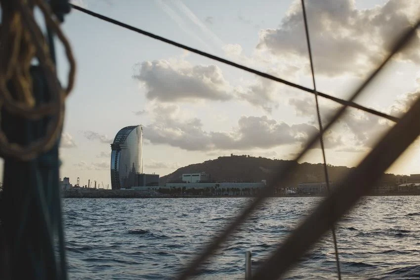 View of Barcelona coastline with the iconic W Hotel building, hills, and water seen from a boat through ropes and rigging.