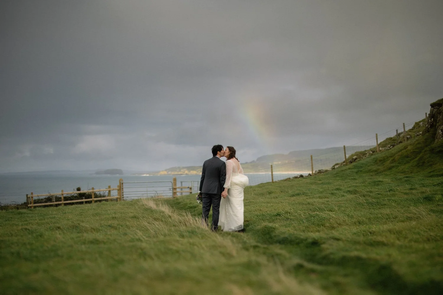 A couple in wedding attire holding hands and kissing on a grassy hill by the ocean. Dark stormy clouds with a faint rainbow in the sky. Fence along the hill's edge.