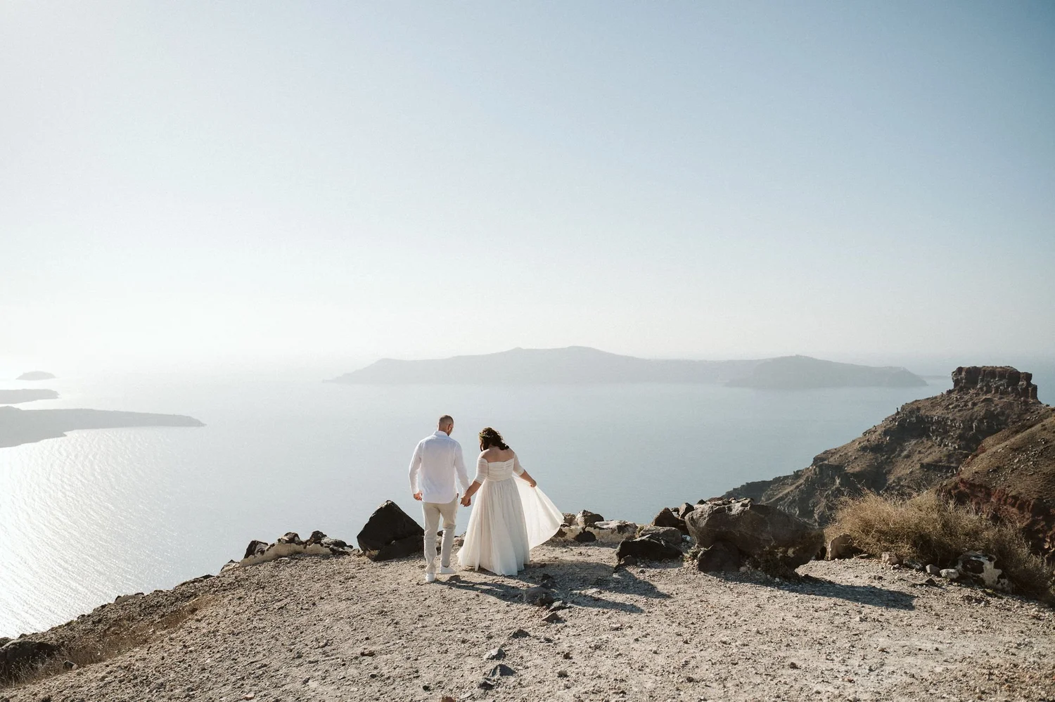 A bride and groom holding hands and walking on rocky terrain overlooking a body of water with islands in the background, clear sky.