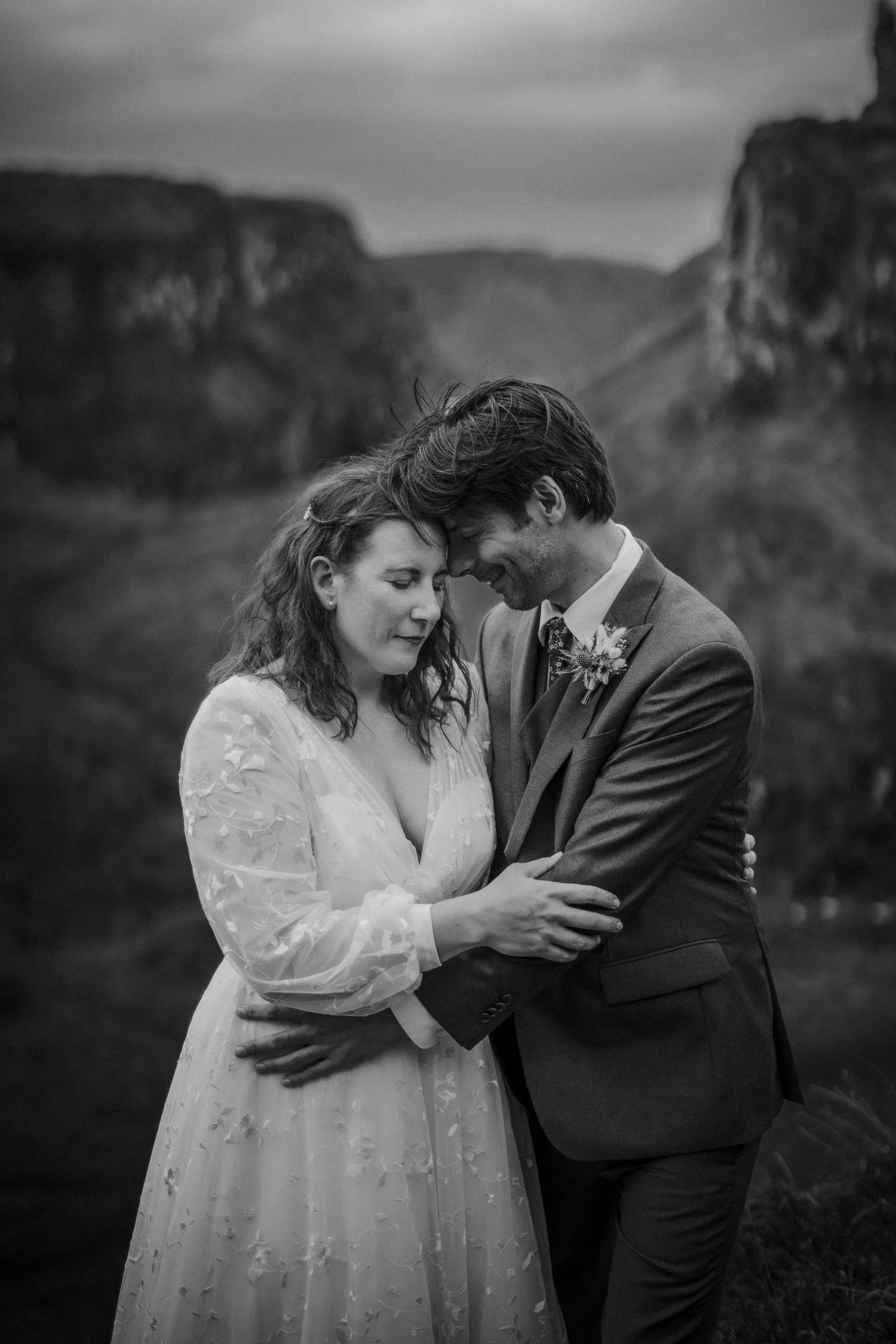 A black and white photo of a couple in wedding attire, standing close and touching foreheads, outdoors with a hilly landscape in the background.