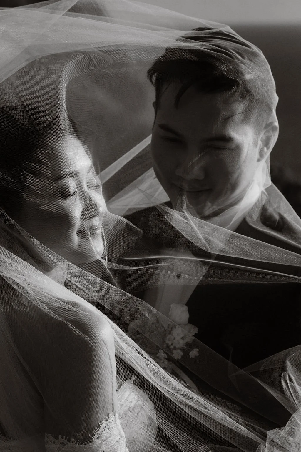 Black and white photo of a smiling bride and groom under a veil, close-up of their faces.