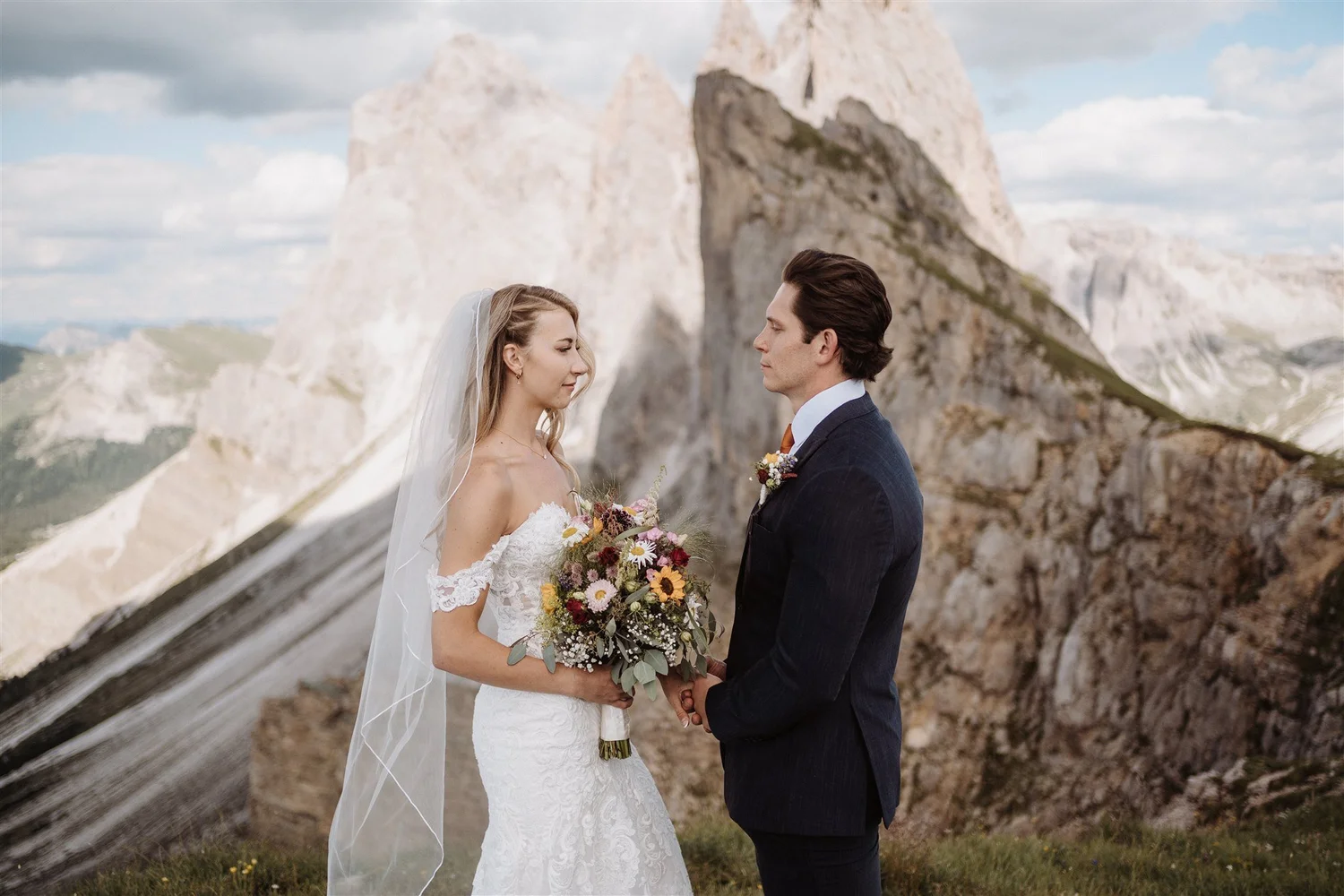 A bride and groom exchanging vows outdoors in front of a mountain landscape with rocky peaks, holding hands, with the bride wearing a white lace wedding dress and veil, and the groom in a dark suit, holding a bouquet of flowers.