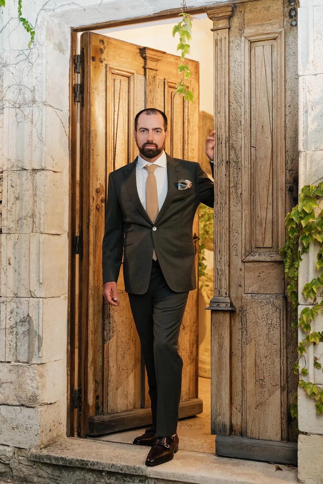 A man in a suit standing in a wooden doorway, looking at the camera, with greenery and ivy around the door frame.
