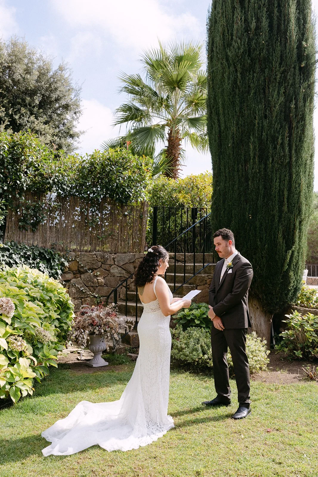 Bride and groom exchanging vows in a garden with lush greenery and tall trees, outdoors during daytime.