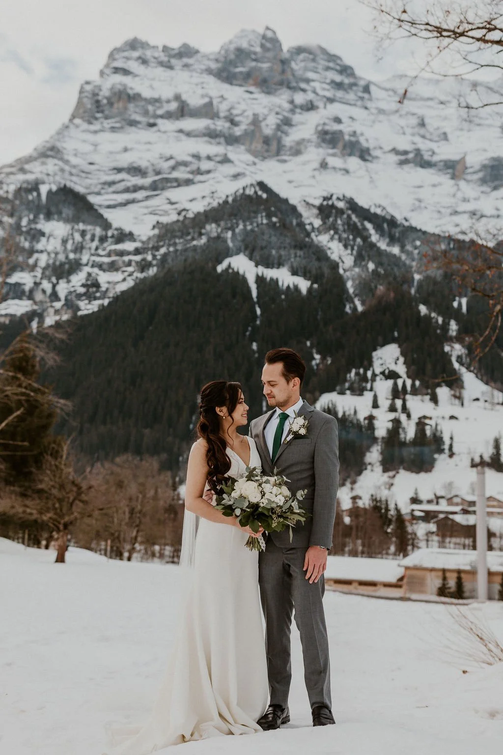 A bride and groom standing in a snowy landscape with a large snow-covered mountain in the background, during winter.
