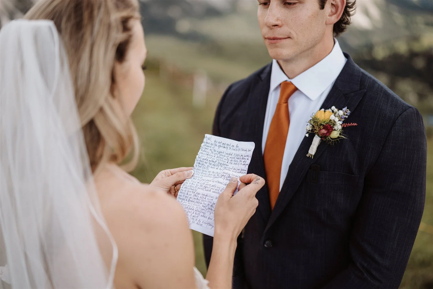 A man in a dark suit with a floral boutonniere is reading a handwritten letter from a woman with long, light-colored hair. They are outdoors with blurred greenery in the background.