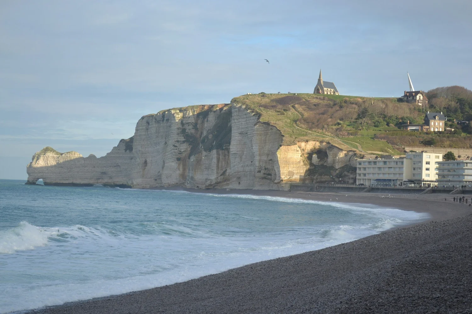 Cliffs near a beach with a church and houses at the top, ocean waves in the foreground, and cloudy sky.