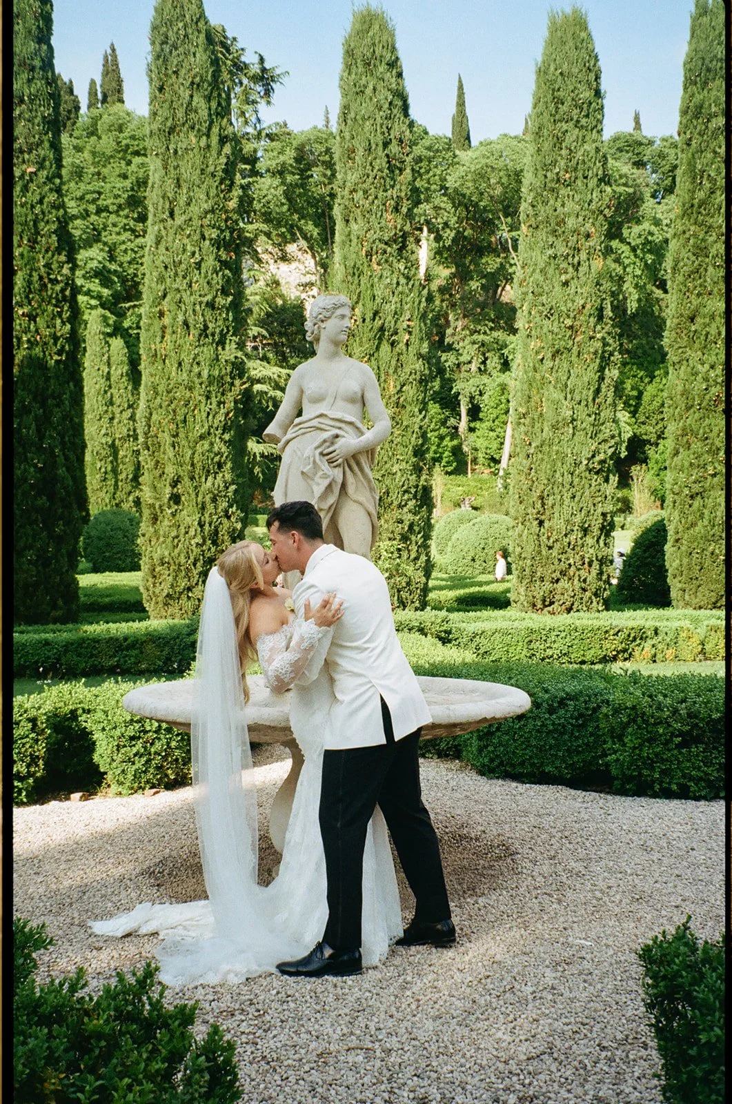 A newlywed couple sharing a kiss in front of a classical marble statue in a lush garden with tall cypress trees.