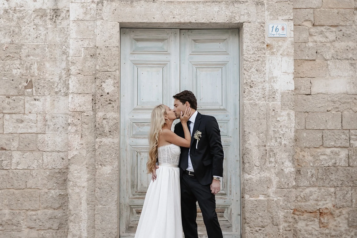 A bride and groom kiss in front of rustic, weathered wooden door set within stone wall, with a street number plaque reading 16 on the wall.