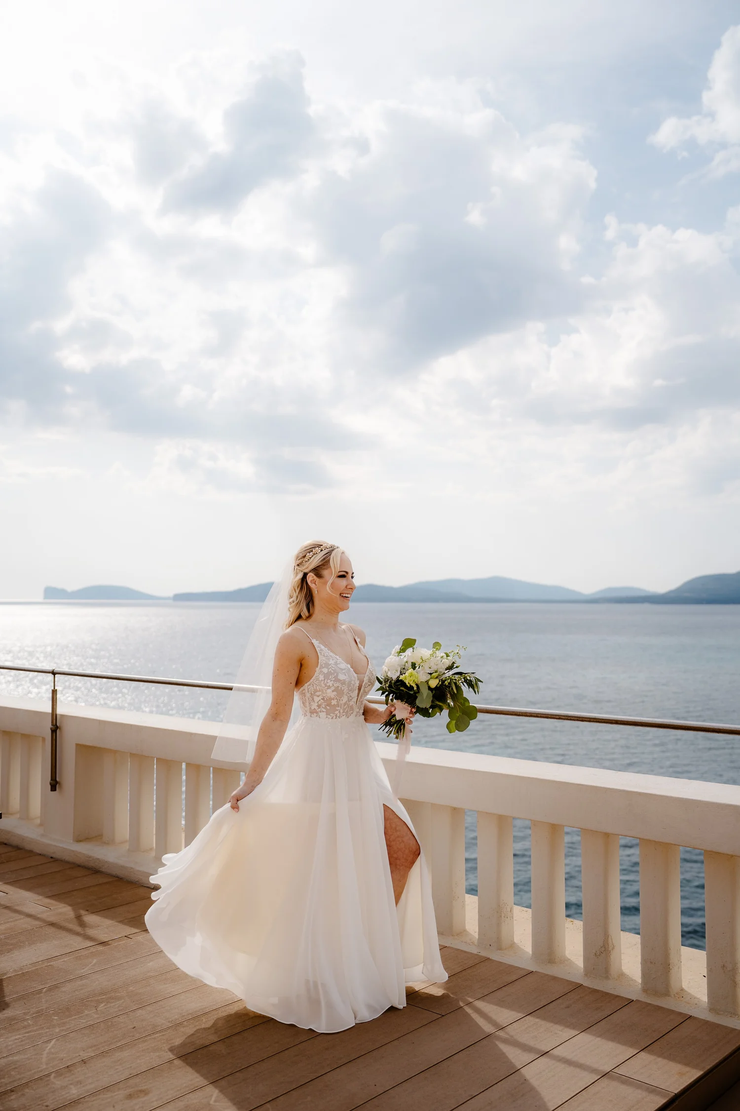 A bride standing on a balcony overlooking the water, holding a bouquet of white flowers with greenery, wearing a white wedding dress with lace detailing and a veil.