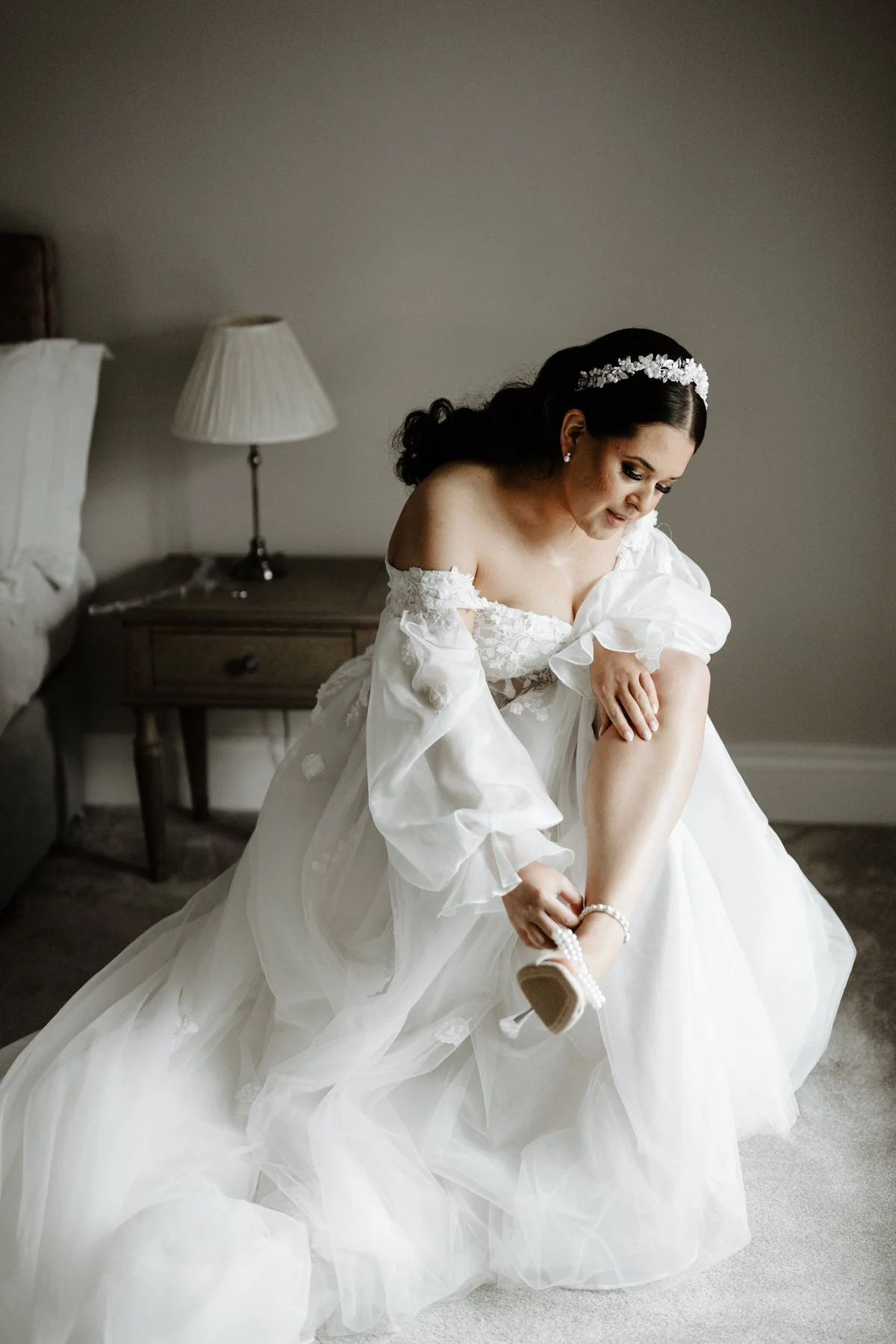 Bridal woman in a wedding gown putting on her shoe in a hotel room.