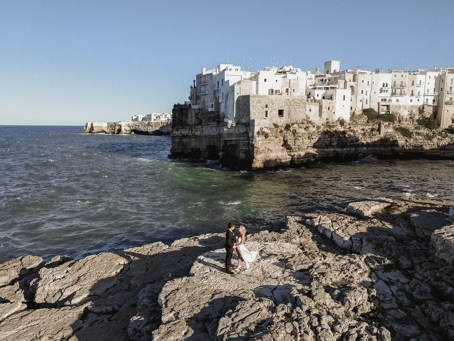 A couple on a rocky shoreline near the sea, with white buildings on a cliff in the background under a clear blue sky.