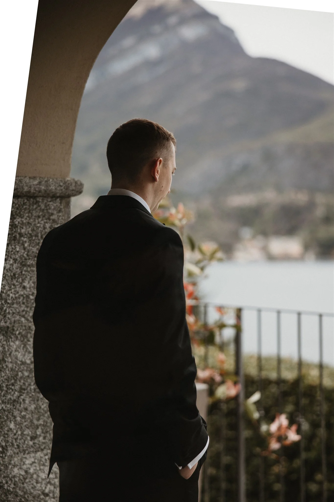 Man in a black suit standing on a balcony, looking at a mountain landscape with a lake and mountains in the distance.