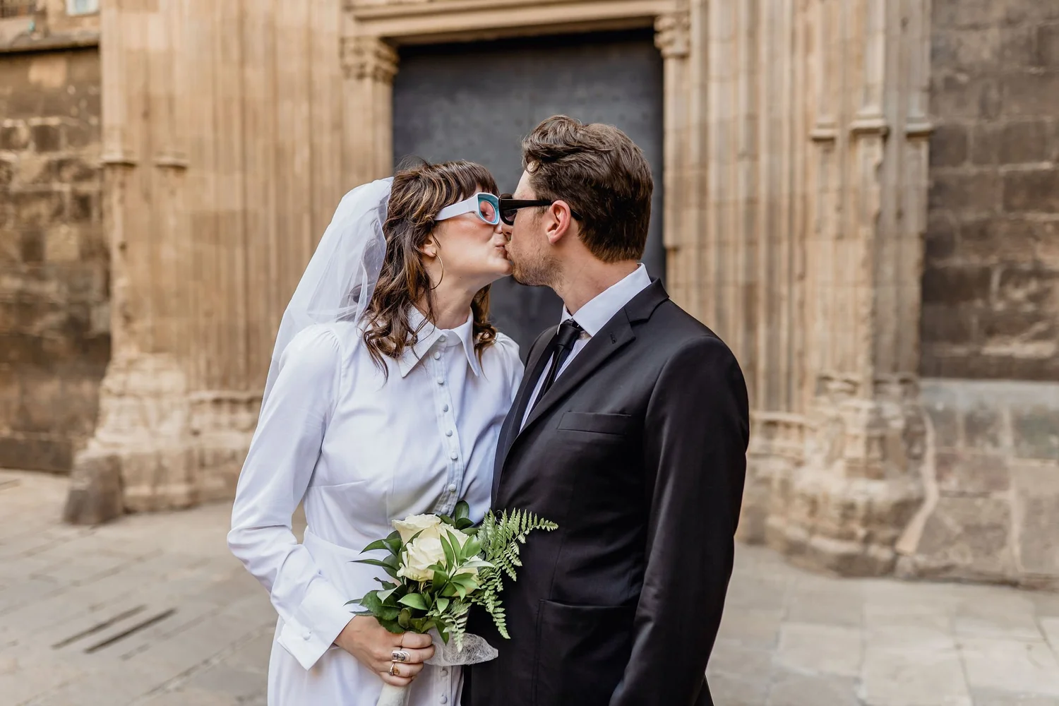 A bride and groom sharing a kiss outside a stone building, with the bride holding a bouquet of white flowers and wearing glasses, and the groom in a formal suit.