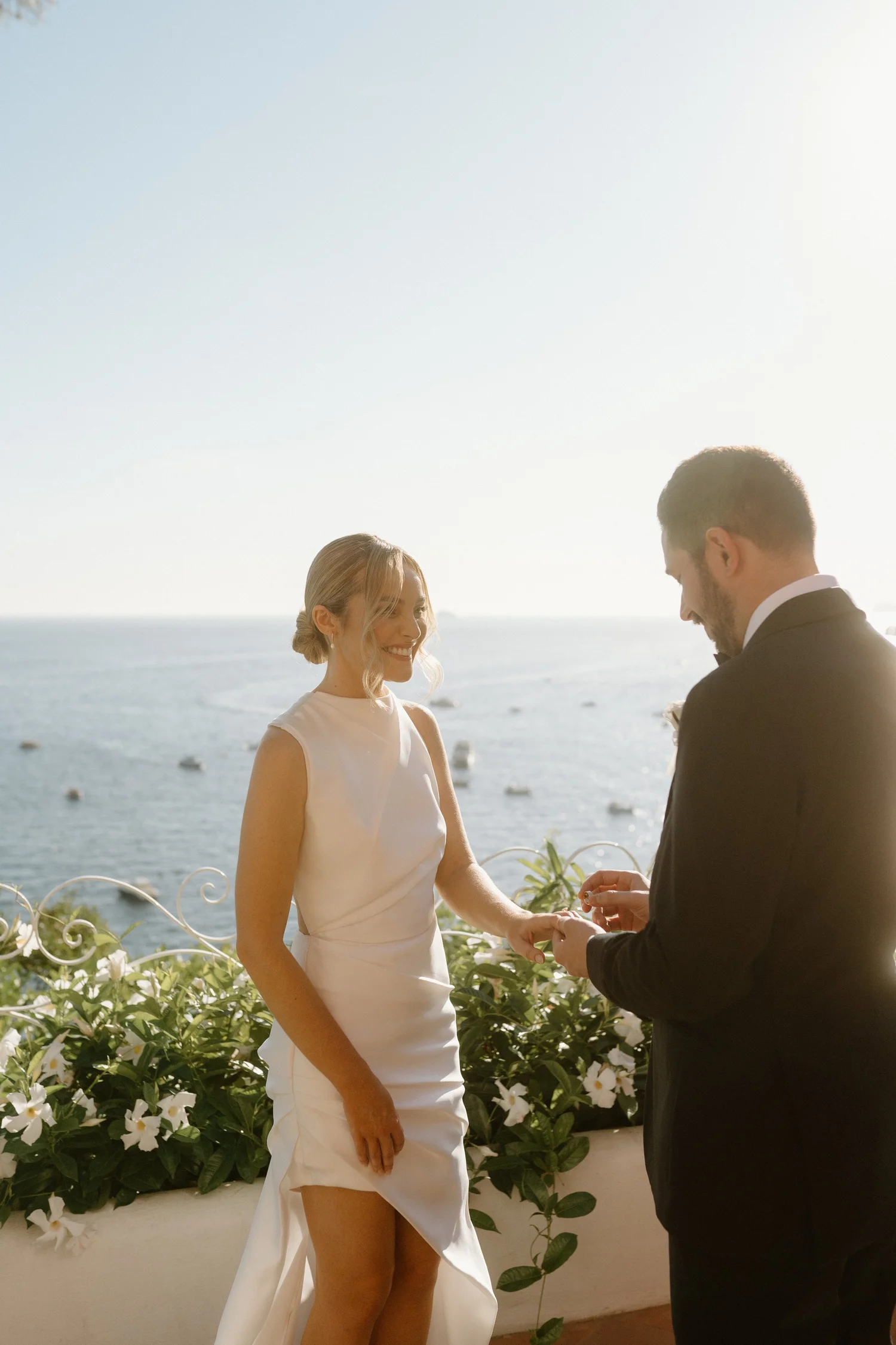 A woman in a white dress and a man in a black suit exchanging rings during a wedding ceremony by the ocean, with white flowers and green foliage in the foreground.