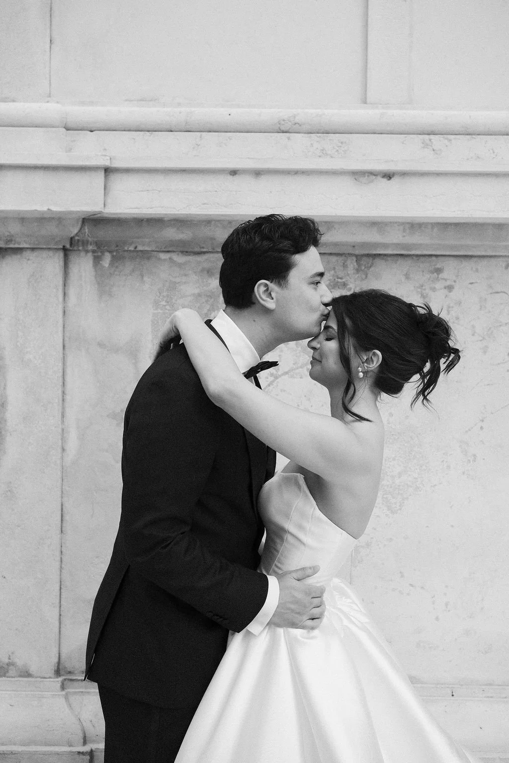 A black and white photo of a bride and groom embracing. The groom is kissing the bride on her forehead, and they are smiling with their eyes closed. The bride has her arms around the groom's neck, and the groom's hands are on her waist. The bride is wearing a strapless wedding dress, and the groom is in a tuxedo.