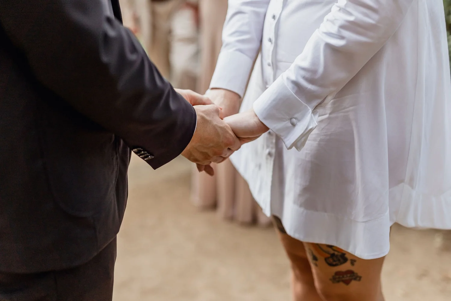 A couple is holding hands during a wedding ceremony, with a woman in a white dress and tattoos on her leg.